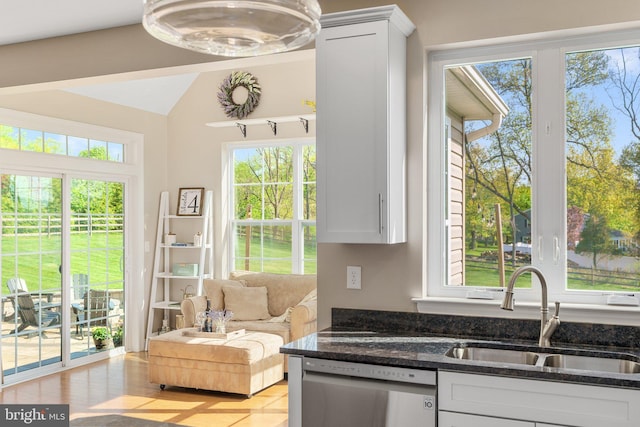 kitchen with stainless steel dishwasher, a sink, white cabinetry, dark stone counters, and vaulted ceiling