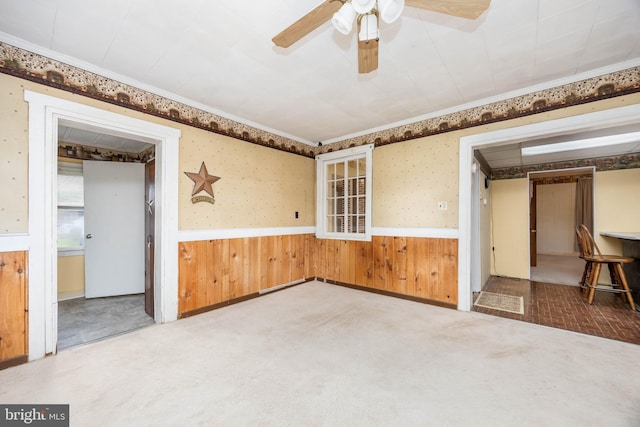 carpeted spare room featuring a wainscoted wall, ceiling fan, wood walls, ornamental molding, and wallpapered walls
