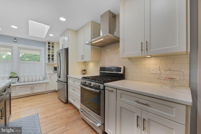 kitchen with stainless steel appliances, wall chimney exhaust hood, a skylight, backsplash, and light wood-type flooring