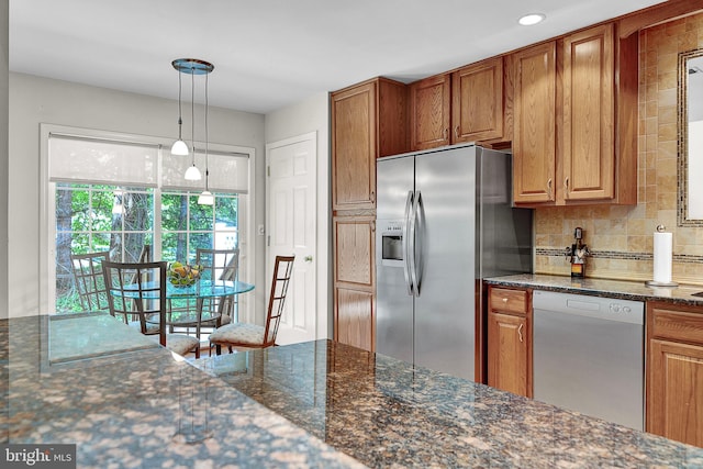 kitchen featuring appliances with stainless steel finishes, decorative backsplash, brown cabinets, hanging light fixtures, and dark stone countertops