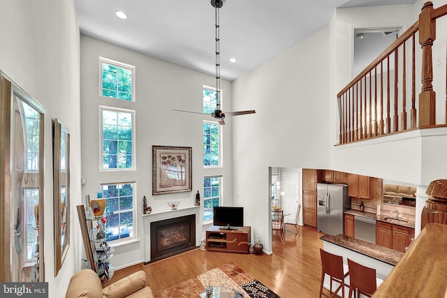 living room with a towering ceiling, a glass covered fireplace, light wood-type flooring, recessed lighting, and a ceiling fan