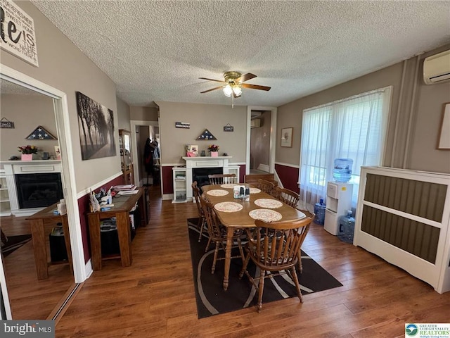 dining room featuring a glass covered fireplace, ceiling fan, a textured ceiling, wood finished floors, and a wall unit AC