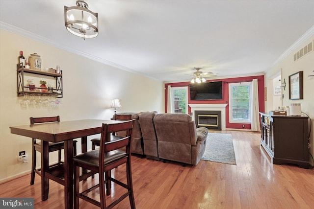 dining area featuring crown molding, light wood-style floors, a glass covered fireplace, a ceiling fan, and baseboards