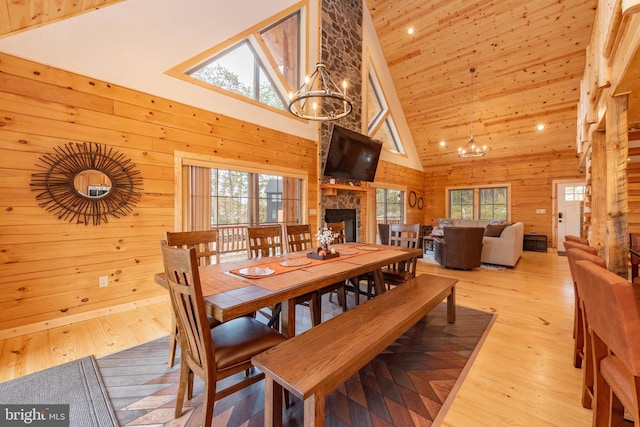 dining area featuring a chandelier, high vaulted ceiling, wooden walls, light wood-type flooring, and a stone fireplace