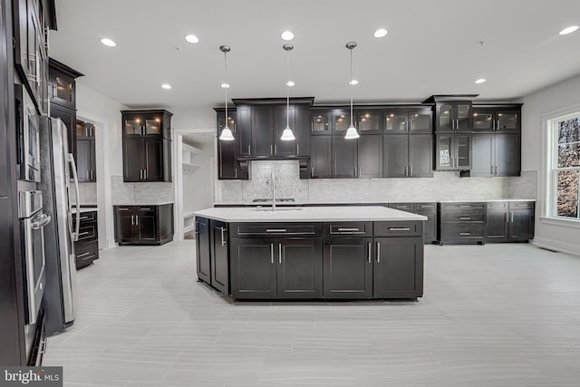 kitchen featuring backsplash, a center island with sink, light countertops, decorative light fixtures, and glass insert cabinets