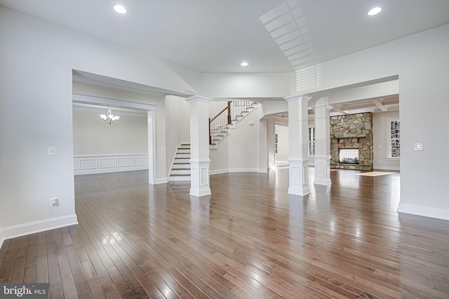 unfurnished living room with hardwood / wood-style floors, stairway, a stone fireplace, decorative columns, and a wainscoted wall