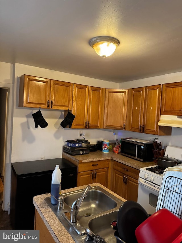 kitchen with under cabinet range hood, white gas range, brown cabinetry, and a sink