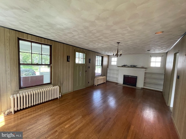 unfurnished living room featuring radiator, wood finished floors, a chandelier, a fireplace with flush hearth, and wood walls