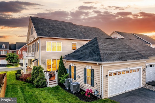 back of house at dusk with roof with shingles, asphalt driveway, a yard, and central air condition unit