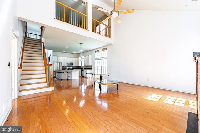 unfurnished living room with a towering ceiling, a ceiling fan, stairs, light wood-type flooring, and baseboards