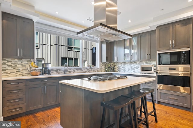 kitchen featuring appliances with stainless steel finishes, island exhaust hood, a sink, light countertops, and decorative backsplash