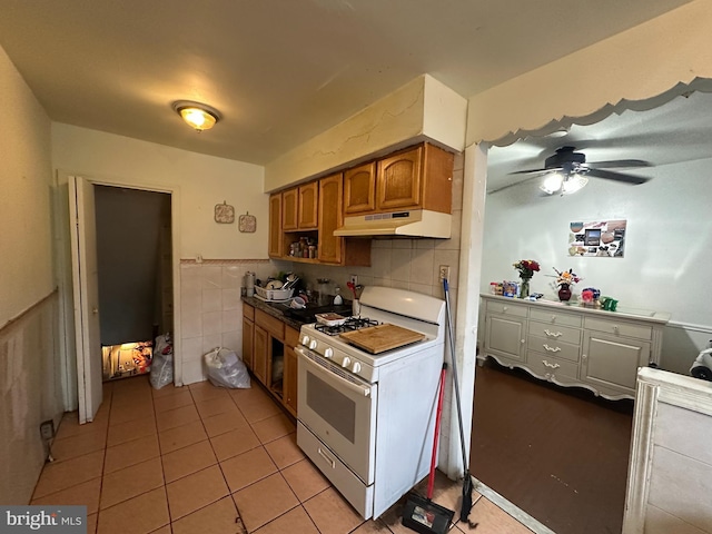 kitchen with white range with gas cooktop, under cabinet range hood, a ceiling fan, light tile patterned flooring, and a wainscoted wall