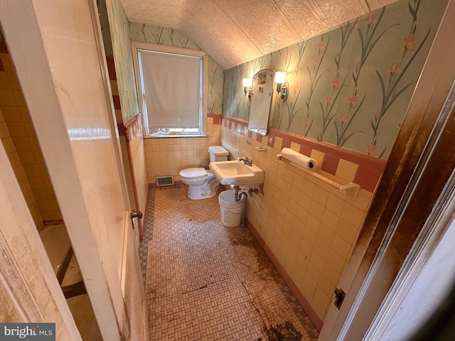bathroom featuring toilet, wallpapered walls, vaulted ceiling, a wainscoted wall, and tile walls