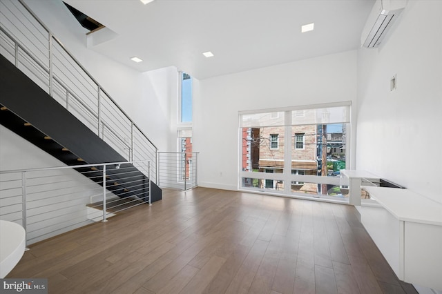 unfurnished living room with a wall unit AC, stairway, wood finished floors, and a towering ceiling