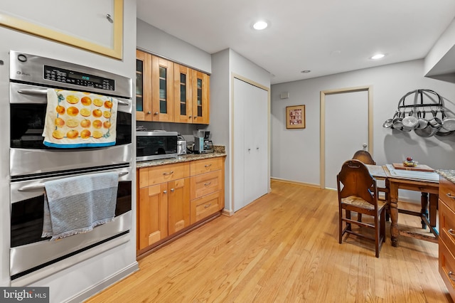 kitchen with appliances with stainless steel finishes, light wood-style floors, glass insert cabinets, light stone countertops, and recessed lighting