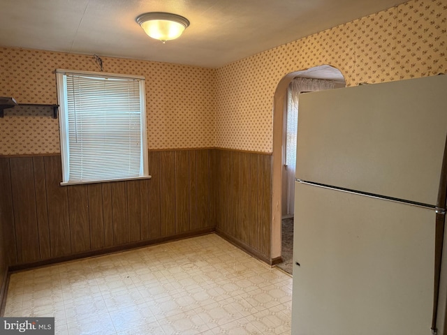 unfurnished dining area with wainscoting, wallpapered walls, light flooring, wooden walls, and arched walkways