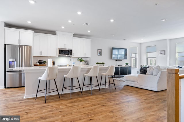 kitchen with appliances with stainless steel finishes, a kitchen island with sink, a kitchen breakfast bar, white cabinets, and light wood-style flooring