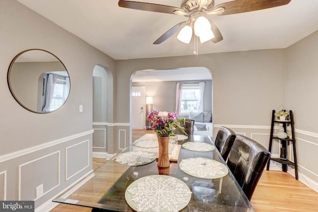 dining room featuring arched walkways, a ceiling fan, a decorative wall, light wood finished floors, and wainscoting