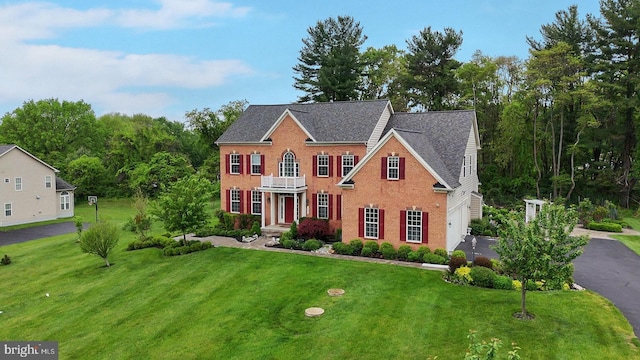 colonial house featuring a front yard, a balcony, a garage, brick siding, and asphalt driveway