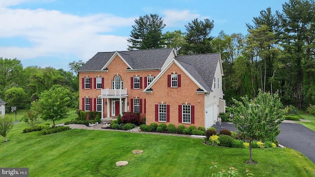 georgian-style home with a balcony, a front yard, a garage, brick siding, and asphalt driveway