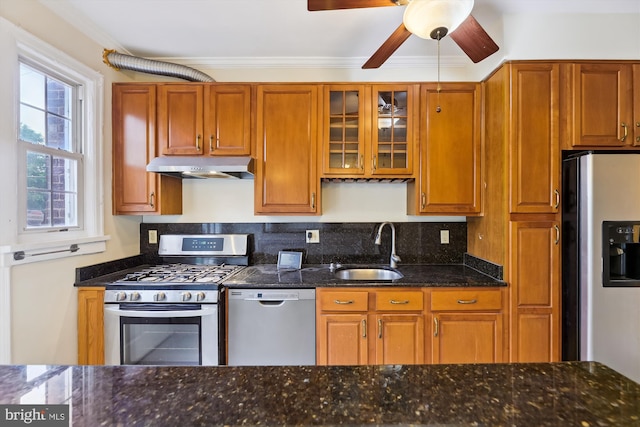 kitchen with appliances with stainless steel finishes, a sink, under cabinet range hood, dark stone counters, and crown molding