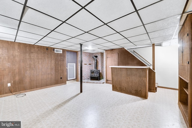 finished below grade area with a wood stove, a paneled ceiling, light flooring, baseboards, and wooden walls