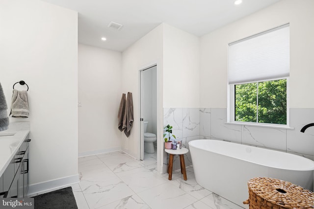 full bath featuring marble look tile flooring, a freestanding tub, toilet, vanity, and a wainscoted wall