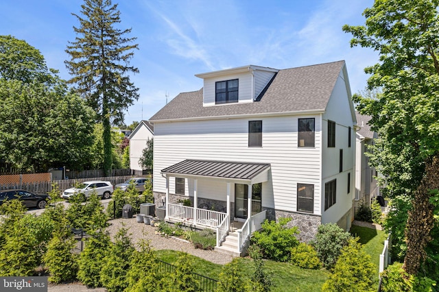 back of house featuring covered porch, stone siding, fence, a shingled roof, and central AC unit