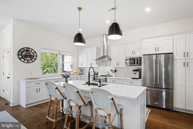 kitchen featuring appliances with stainless steel finishes, wall chimney exhaust hood, a sink, dark wood-style flooring, and an island with sink