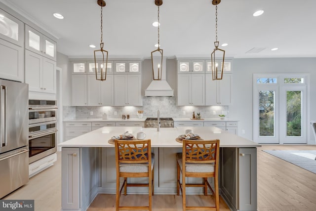 kitchen with appliances with stainless steel finishes, premium range hood, backsplash, light countertops, and a center island with sink