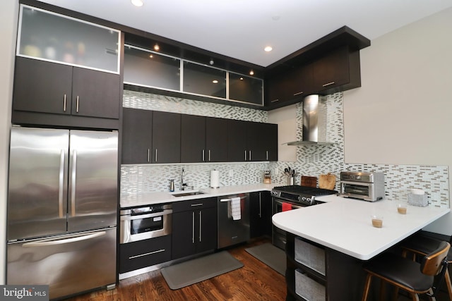 kitchen with stainless steel appliances, wall chimney range hood, a peninsula, a sink, and dark cabinetry