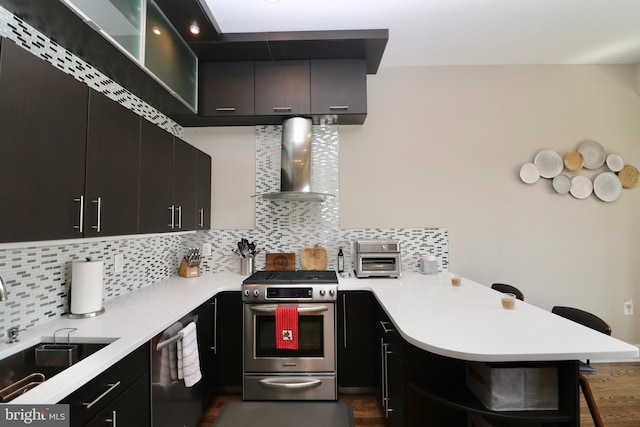 kitchen featuring wall chimney range hood, stainless steel appliances, a peninsula, a sink, and a breakfast bar area