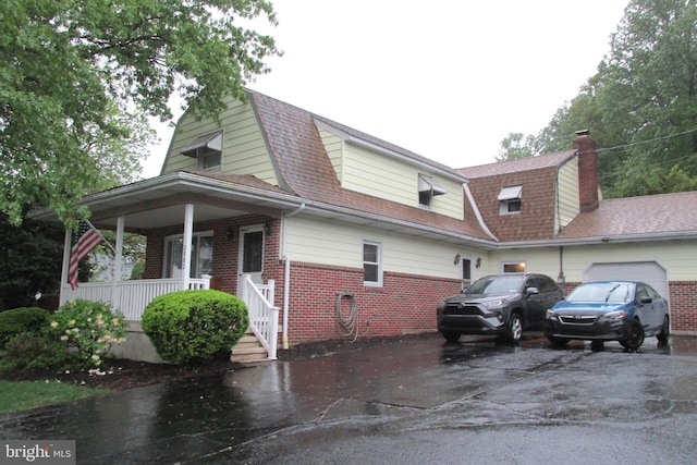 view of home's exterior featuring brick siding, driveway, a gambrel roof, and covered porch