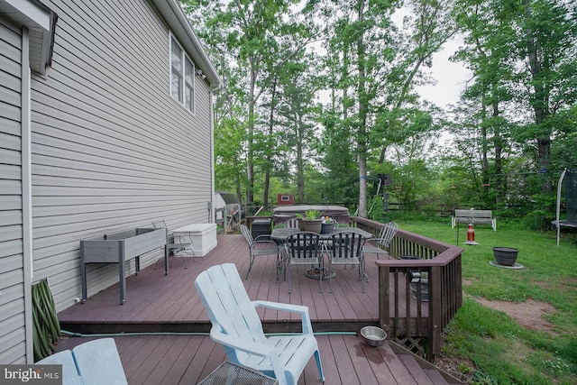 deck with a trampoline, outdoor dining space, a yard, and view of scattered trees