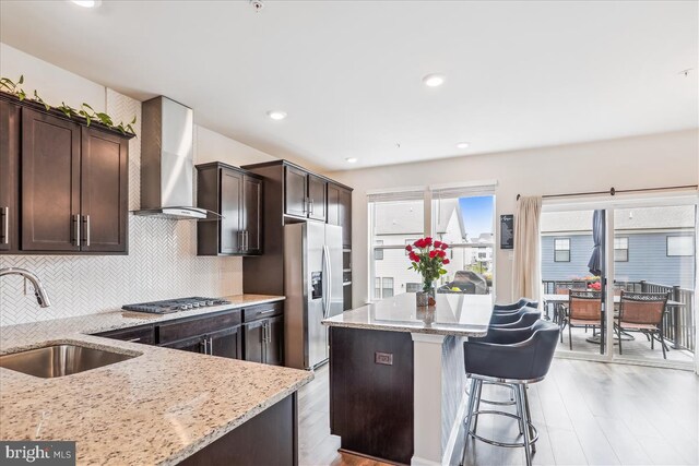 kitchen featuring wall chimney exhaust hood, stainless steel appliances, a sink, light stone counters, and a kitchen island
