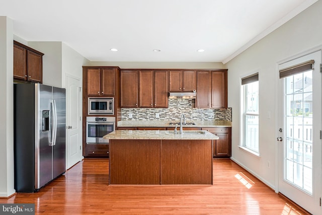 kitchen with stainless steel appliances, under cabinet range hood, tasteful backsplash, light wood-style flooring, and an island with sink