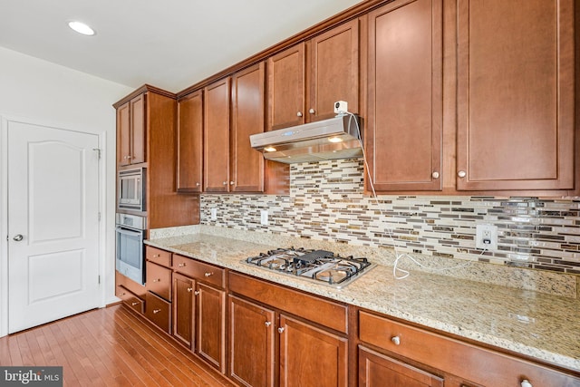 kitchen with appliances with stainless steel finishes, under cabinet range hood, brown cabinetry, light stone counters, and wood finished floors