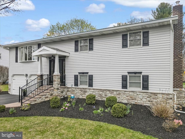 raised ranch with a garage, stone siding, a chimney, and asphalt driveway