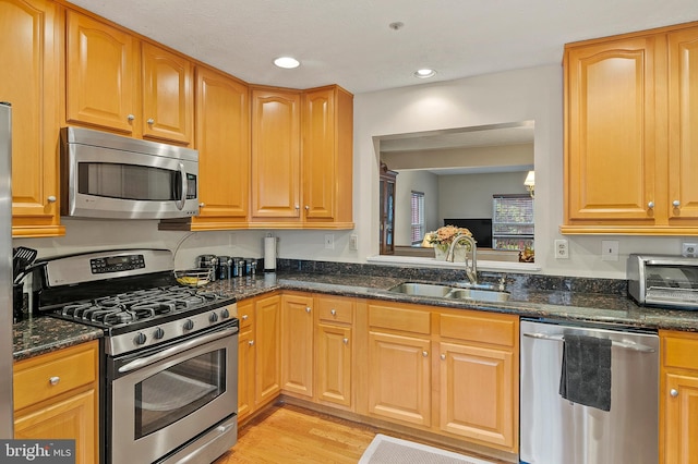 kitchen featuring appliances with stainless steel finishes, a sink, dark stone counters, light wood-type flooring, and recessed lighting