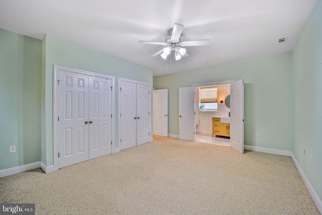 unfurnished bedroom featuring light colored carpet, multiple closets, baseboards, ensuite bath, and ceiling fan