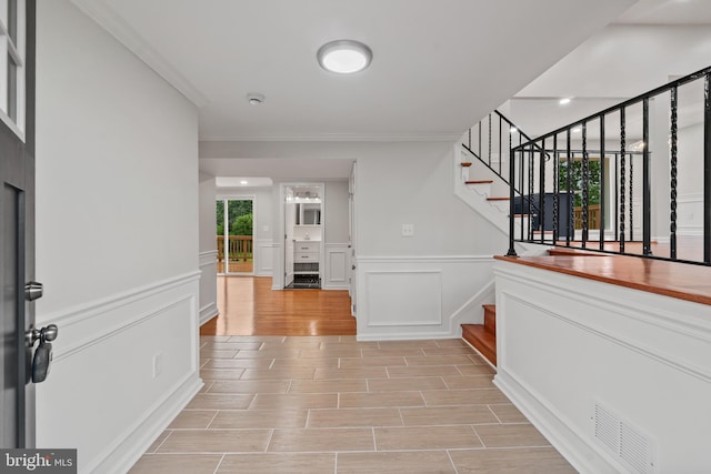 hall with stairs, light wood-style floors, ornamental molding, a decorative wall, and wainscoting
