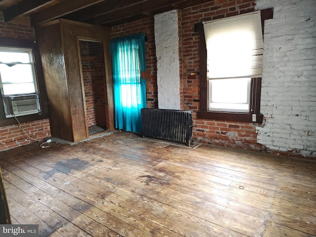 unfurnished living room featuring hardwood / wood-style flooring, radiator heating unit, plenty of natural light, beamed ceiling, and brick wall