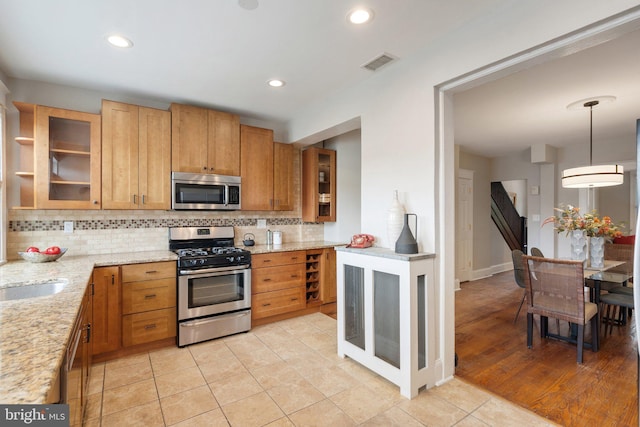 kitchen with appliances with stainless steel finishes, decorative backsplash, light stone countertops, glass insert cabinets, and recessed lighting