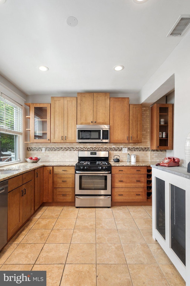kitchen featuring appliances with stainless steel finishes, decorative backsplash, glass insert cabinets, light stone countertops, and recessed lighting