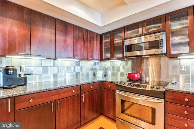kitchen featuring appliances with stainless steel finishes, tasteful backsplash, light stone countertops, glass insert cabinets, and light wood-type flooring