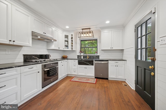kitchen with appliances with stainless steel finishes, under cabinet range hood, dark countertops, hardwood / wood-style floors, and white cabinetry