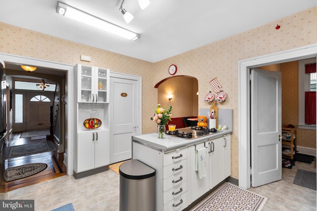 kitchen featuring wallpapered walls, stainless steel gas stovetop, white cabinetry, a peninsula, and glass insert cabinets