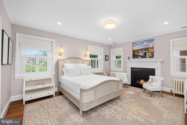bedroom with a warm lit fireplace, radiator, wood finished floors, baseboards, and recessed lighting