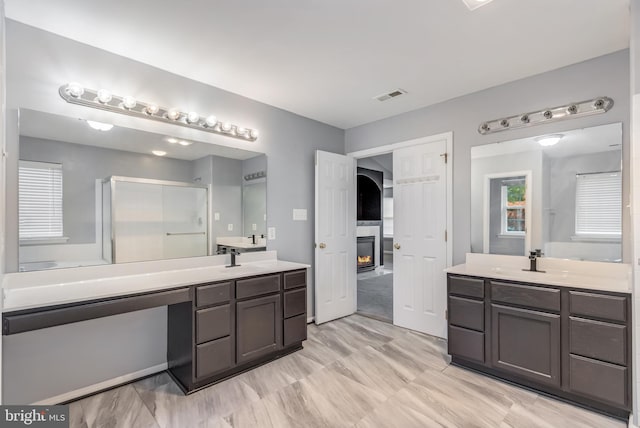 full bathroom featuring a glass covered fireplace, two vanities, and walk in shower