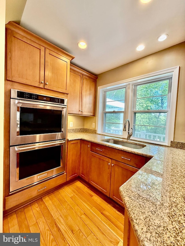 kitchen with a sink, stainless steel double oven, light wood-style flooring, light stone countertops, and recessed lighting